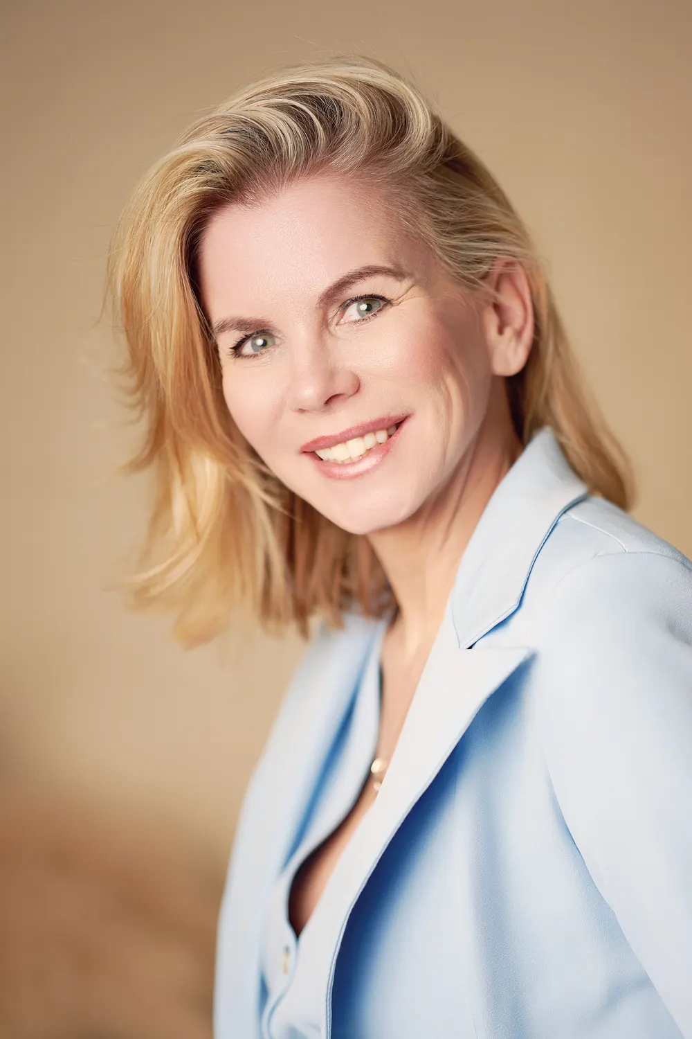 Smiling business portrait of a blonde woman in a light blue blazer against a warm studio background.